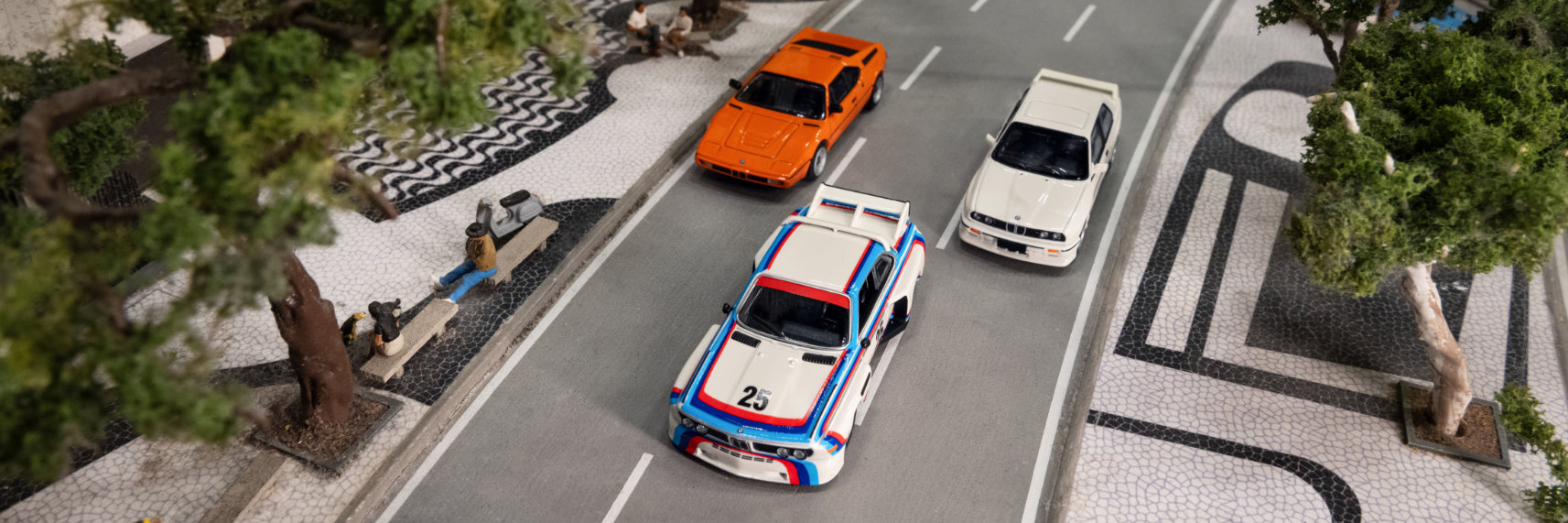 3 BMW miniature cars sit on a play street next to model park benches and trees.