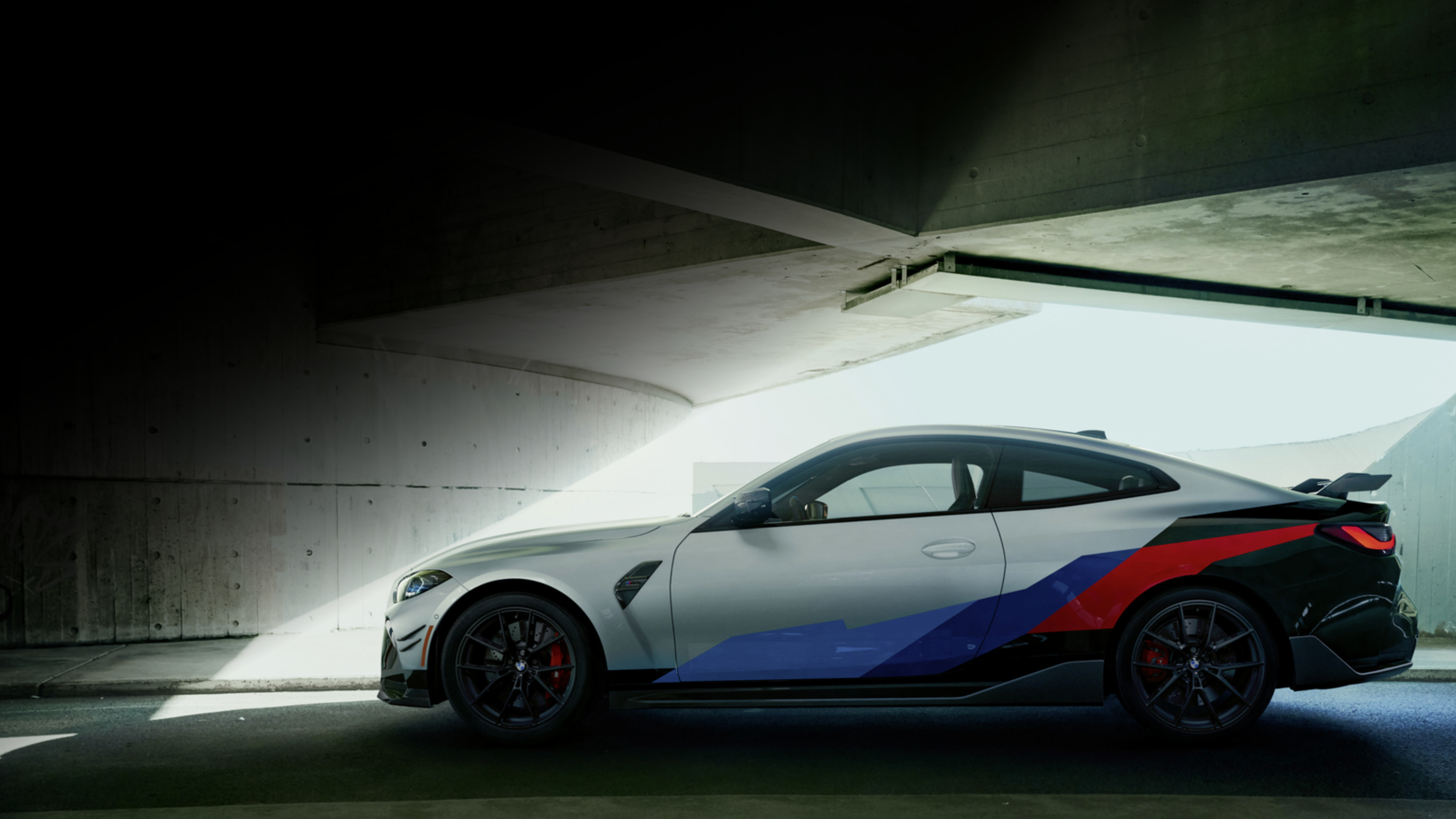 A BMW M4 Coupe with racing stripes sits under a bridge.