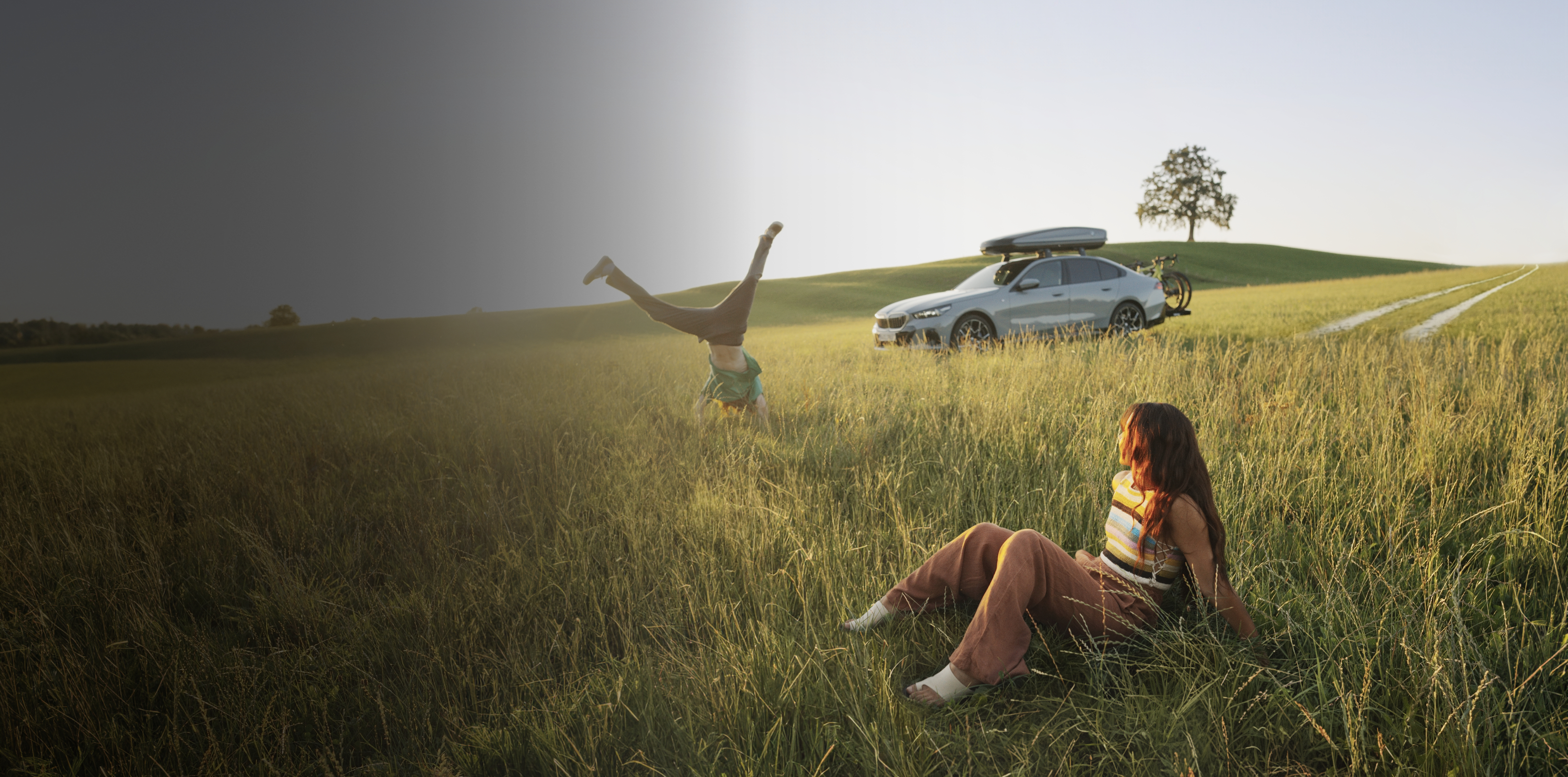 A BMW Sedan with a roof rack is parked in a field with a young woman admiring it over her shoulder in the foreground.