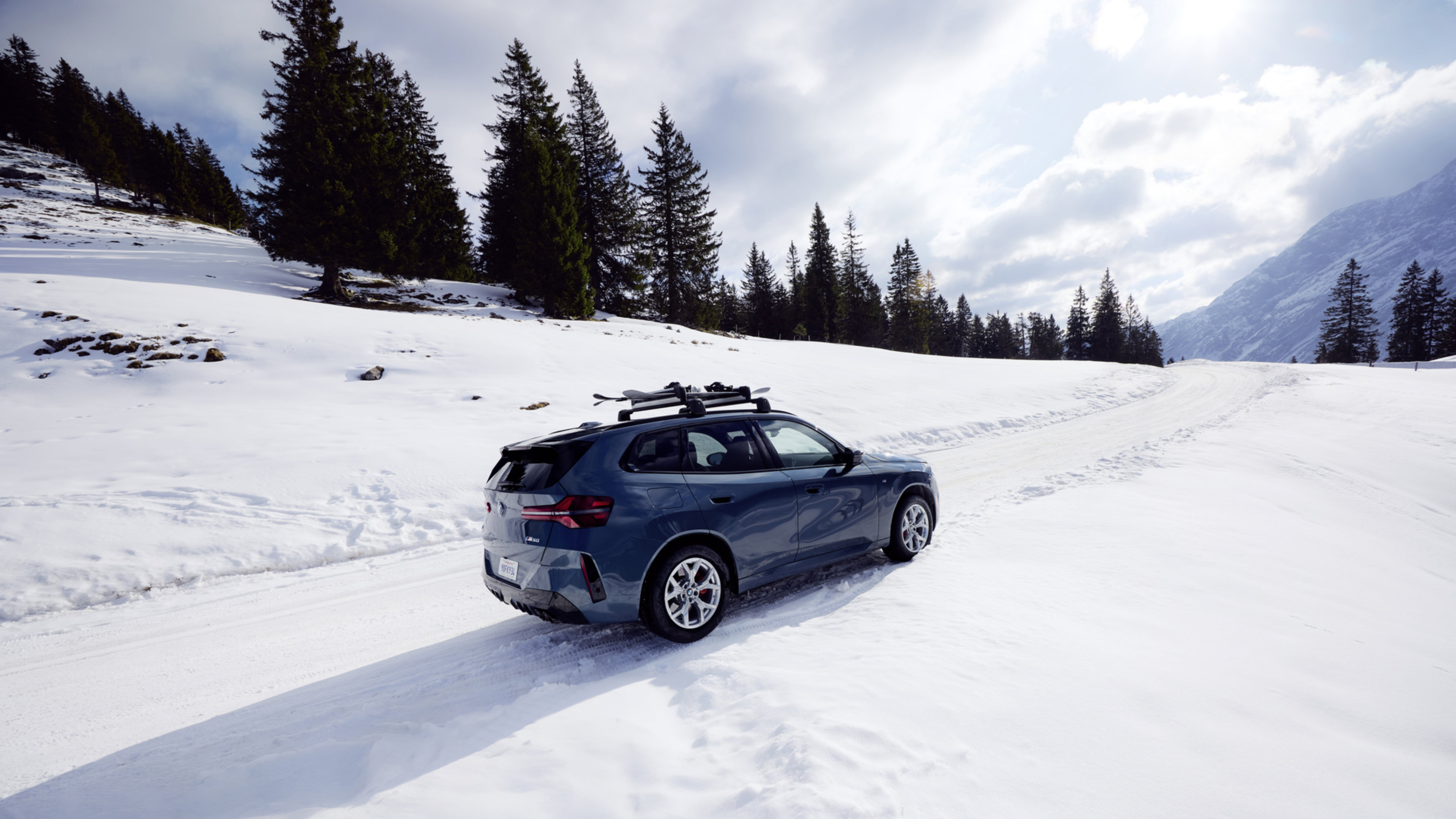 A blue BMW X3 M50i with a BMW roof rack climbs a snowy terrain with pine trees and clouds and sky in the background.